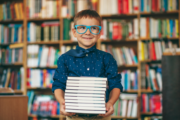Front view, smiling pupil with blue glasses frame and blue tie on a book background. Boy holding a stack of books in his hands against multi colored bookshelf in library. 